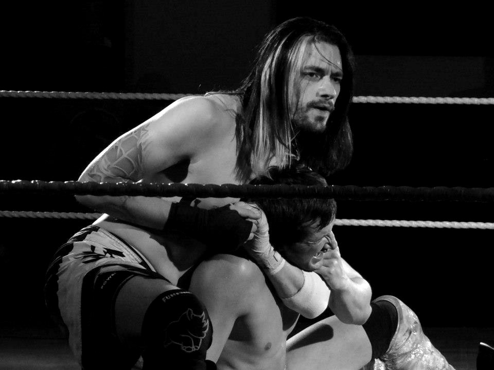 The author mid-match, long hair over one shoulder, locking a chinlock on a seated opponent between the ropes